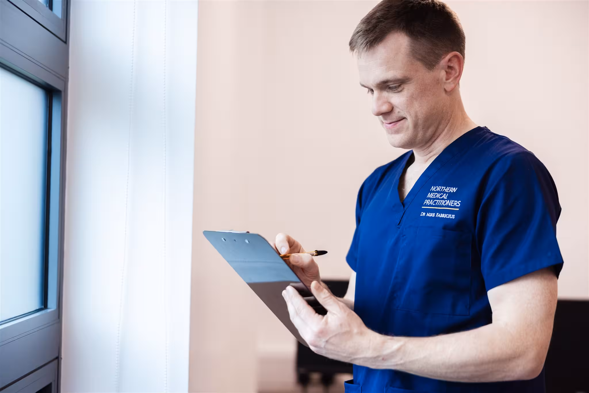 Dr Mike Fabricius of Northern Medical Practitioners in blue scrubs, smiling whilst writing notes on a clipboard.