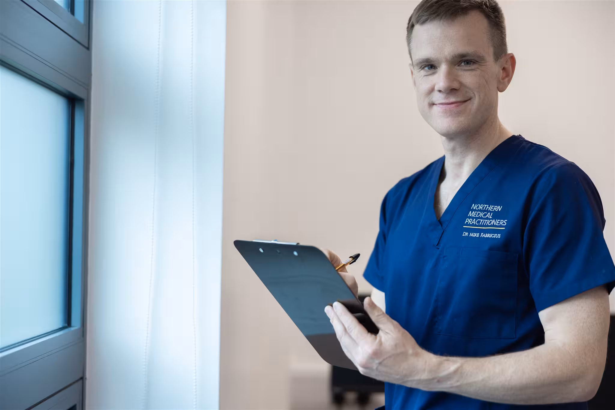 Dr Mike Fabricius of Northern Medical Practitioners, wearing blue scrubs, smiling at the camera whilst holding a clipboard near a bright window.