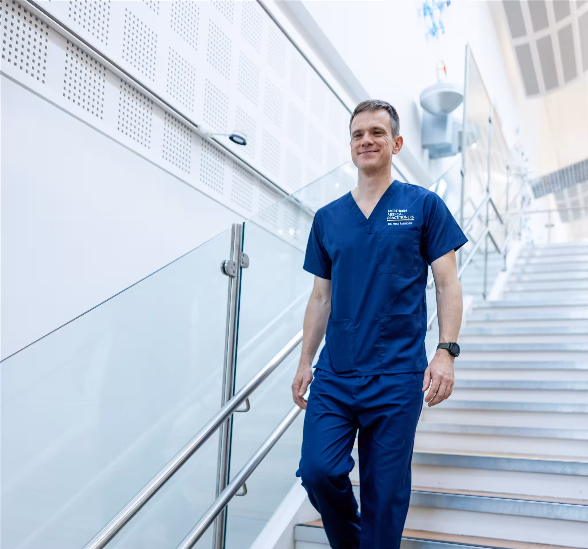 Dr Mike Fabricius of Northern Medical Practitioners, wearing blue scrubs, smiling as he walks down a modern glass-panelled staircase.