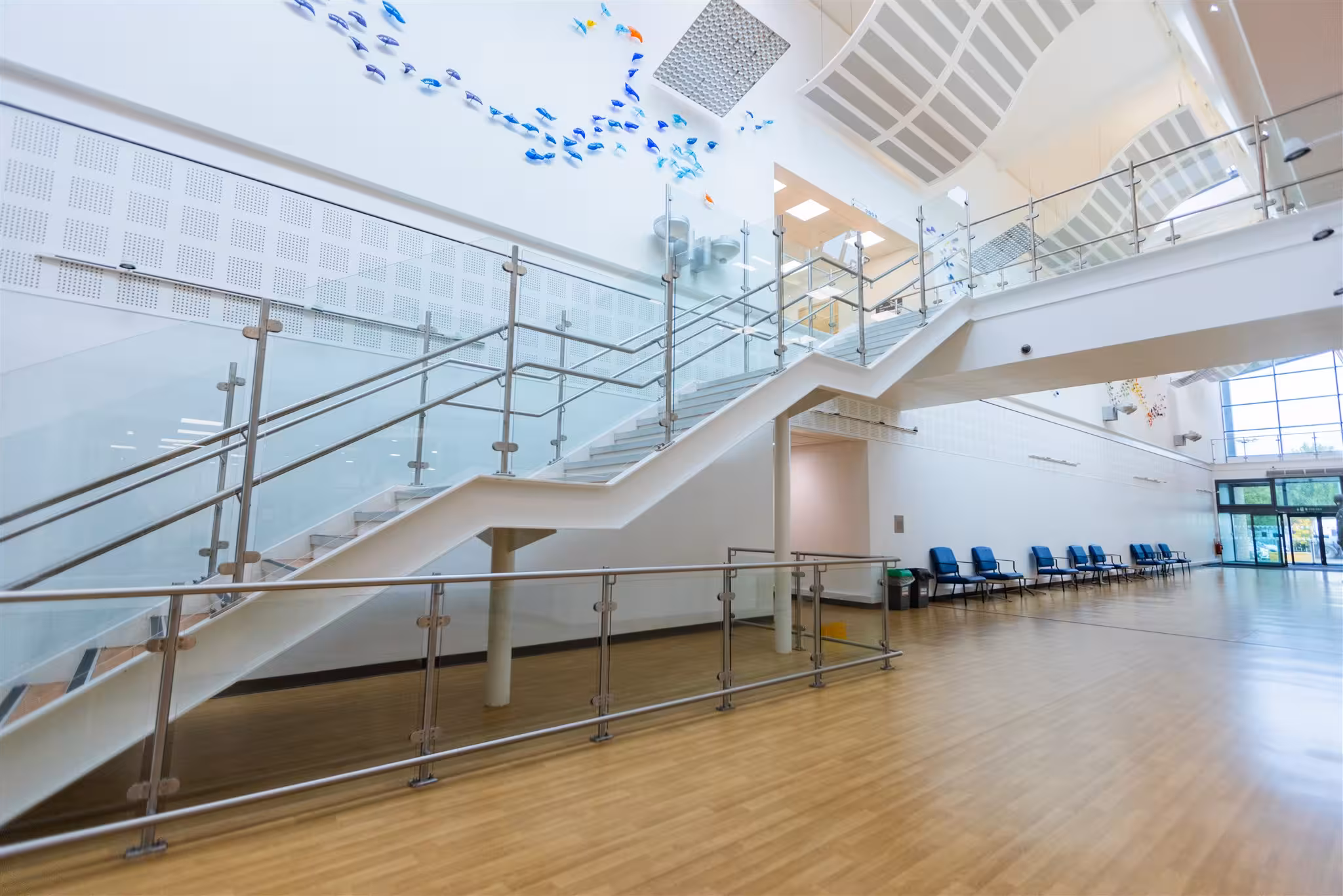 Bright, spacious atrium of a modern healthcare centre featuring a prominent glass-panelled staircase, wooden flooring, and a row of blue waiting chairs.