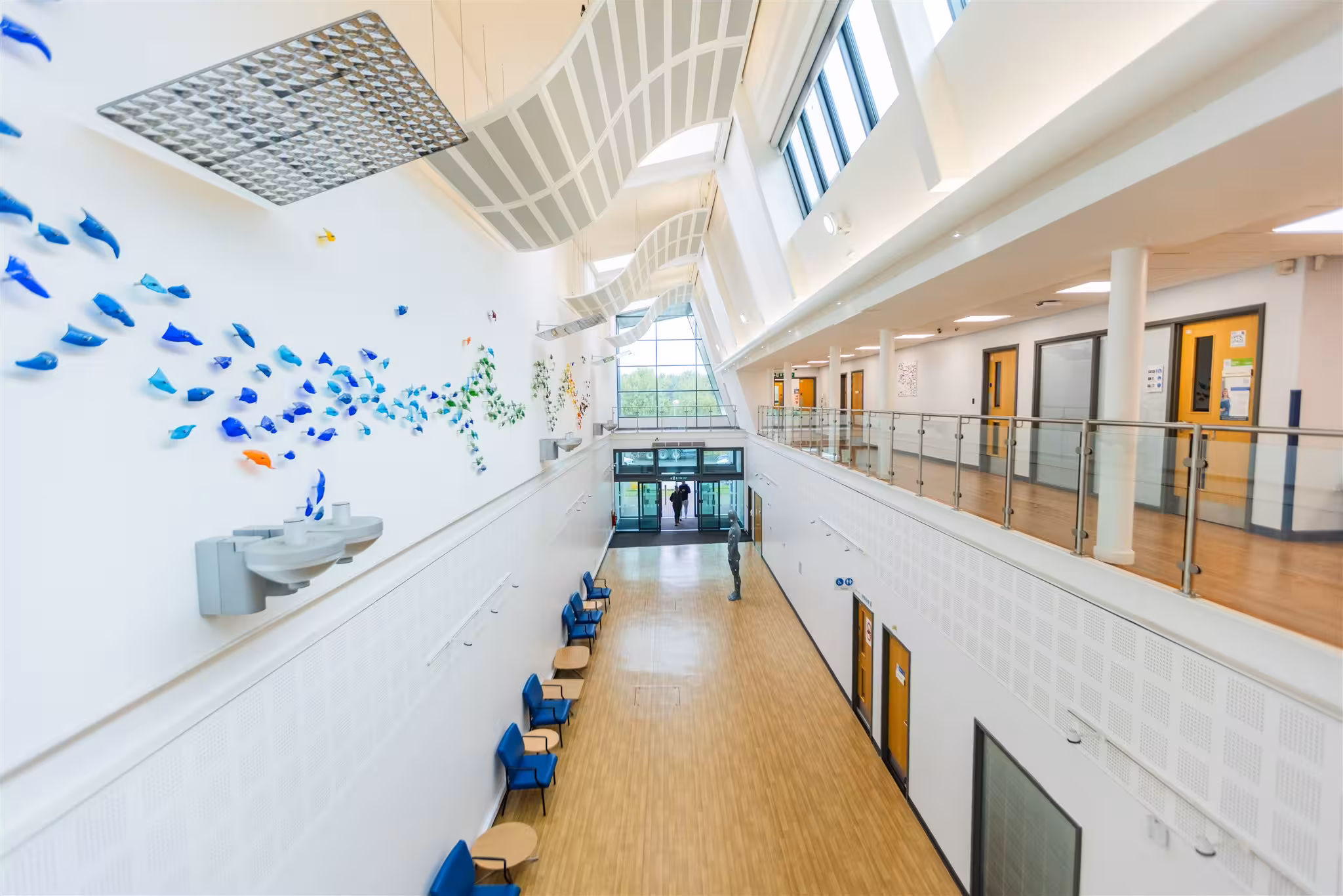 High-angle view of a bright, spacious clinic atrium featuring wooden flooring, a glass-panelled upper walkway, blue waiting chairs, and a decorative blue glass art installation on the wall.