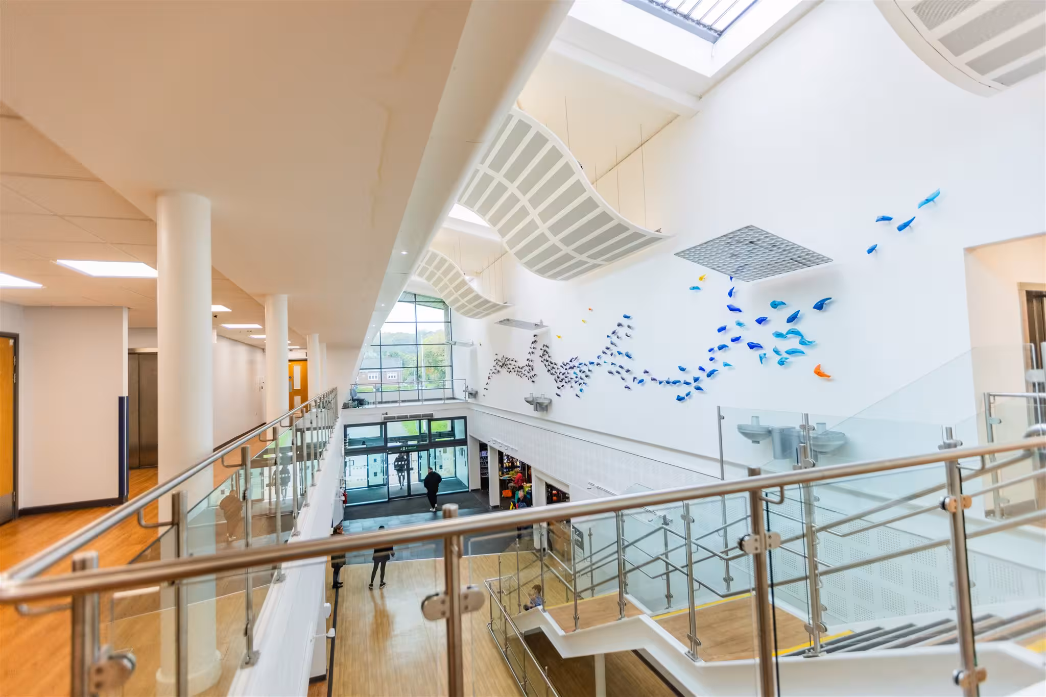 High-angle view of a bright, spacious healthcare centre atrium featuring glass-panelled walkways, wooden flooring, the main entrance doors, and a decorative blue glass art installation on a large white wall.