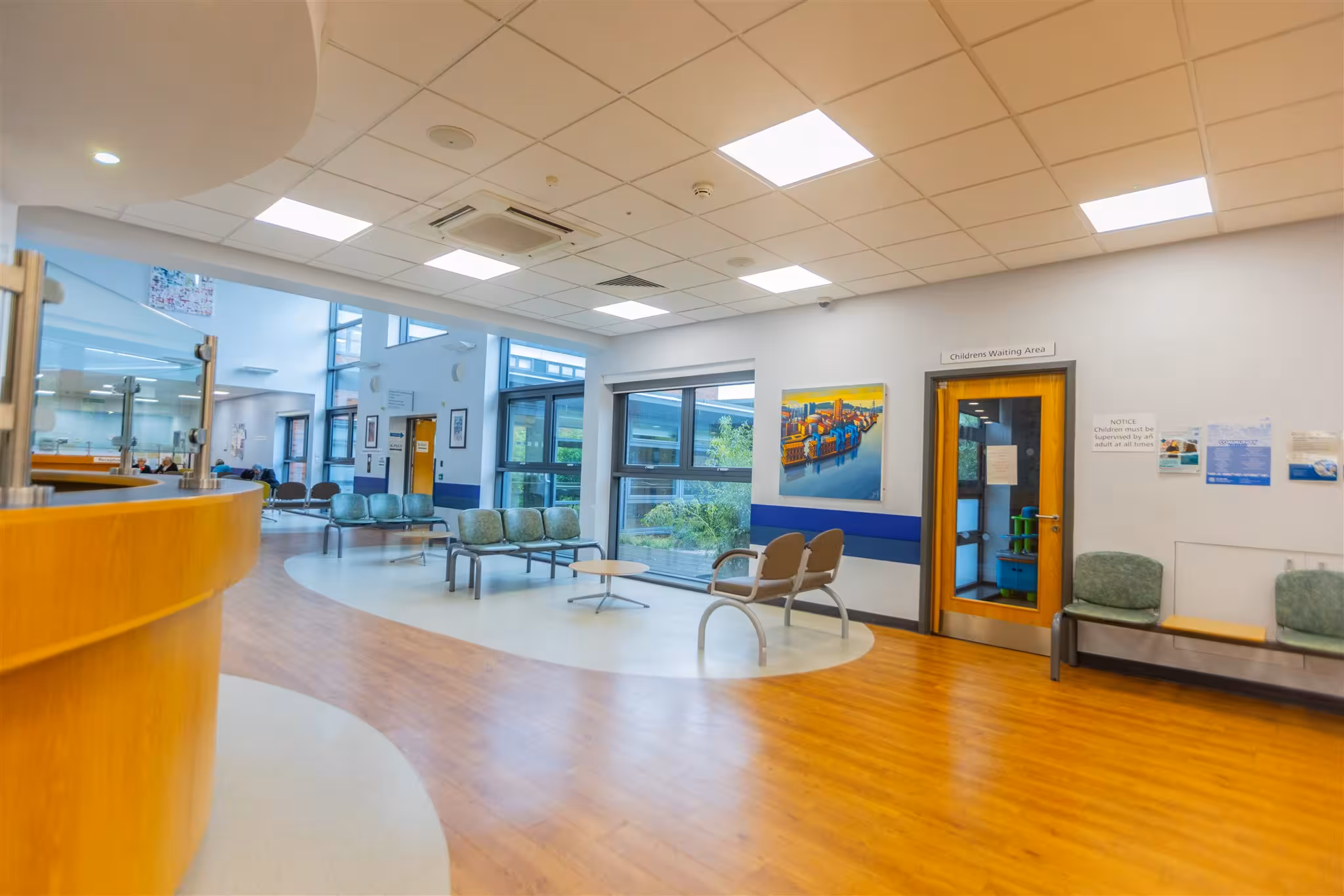A bright, modern clinic waiting area featuring a curved wooden reception desk, scattered green and brown seating, large windows, and a dedicated room labelled 'Childrens Waiting Area'.