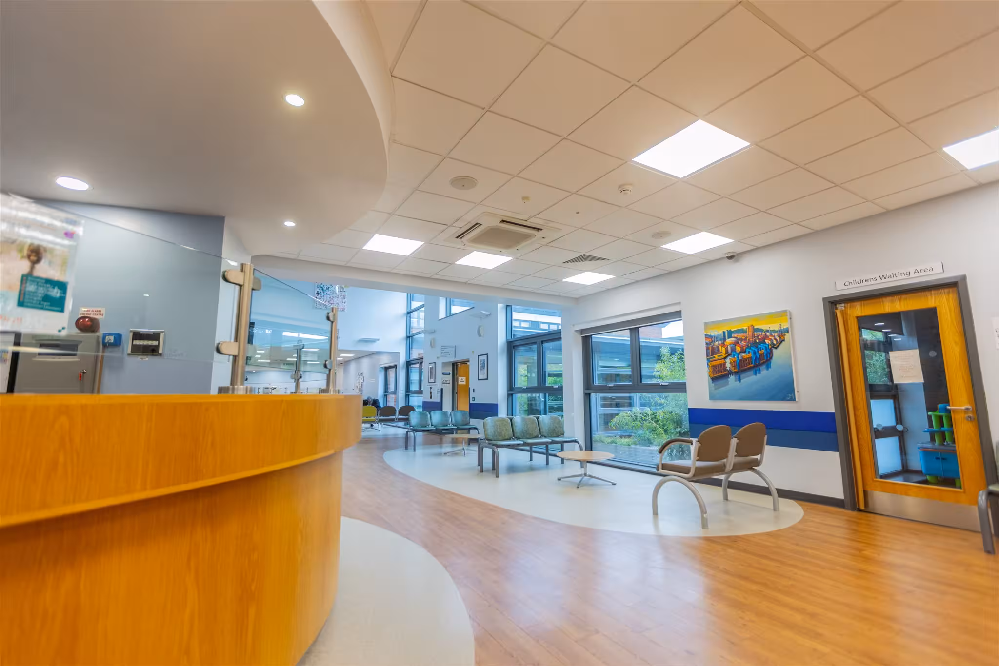Modern and welcoming waiting area of a Northern Medical Practitioners clinic, featuring a curved wooden reception desk, various seating options, large windows, and a dedicated door marked 'Children's Waiting Area'.