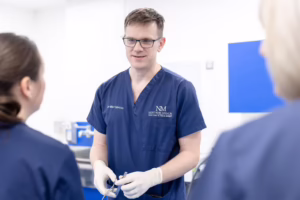 Dr Mike Fabricius of Northern Medical Practitioners, wearing blue scrubs and surgical gloves, holding medical instruments whilst conversing with colleagues in a clinic room.