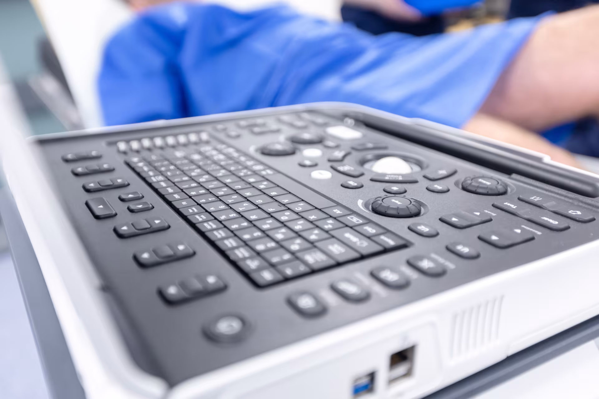 Close-up of the control panel and keyboard of a medical ultrasound machine, with a patient lying on an examination couch in the blurred background.