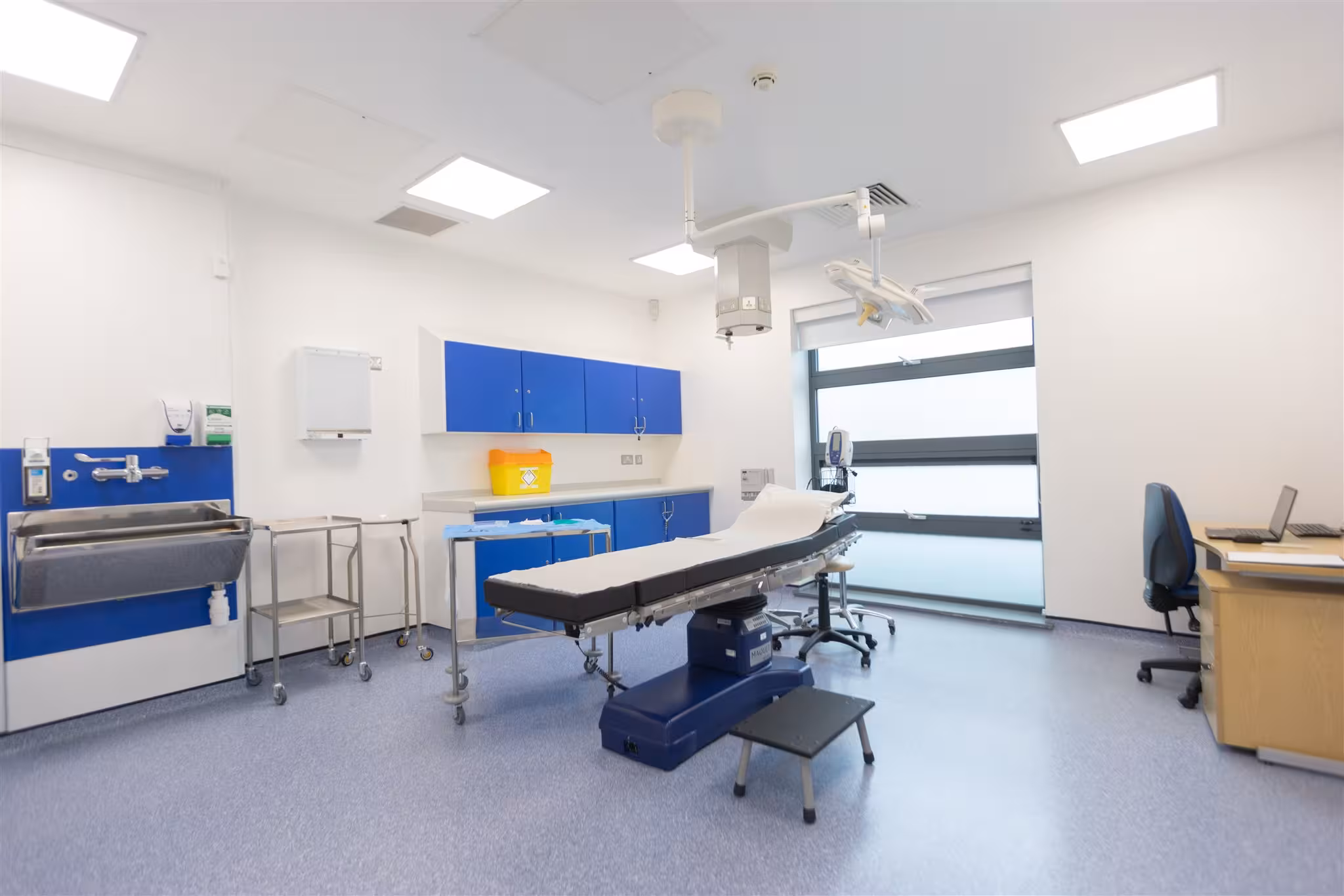 A bright, modern minor surgery and treatment room featuring a central adjustable patient couch, overhead surgical lighting, blue cabinetry, a stainless steel scrub sink, and a desk area.