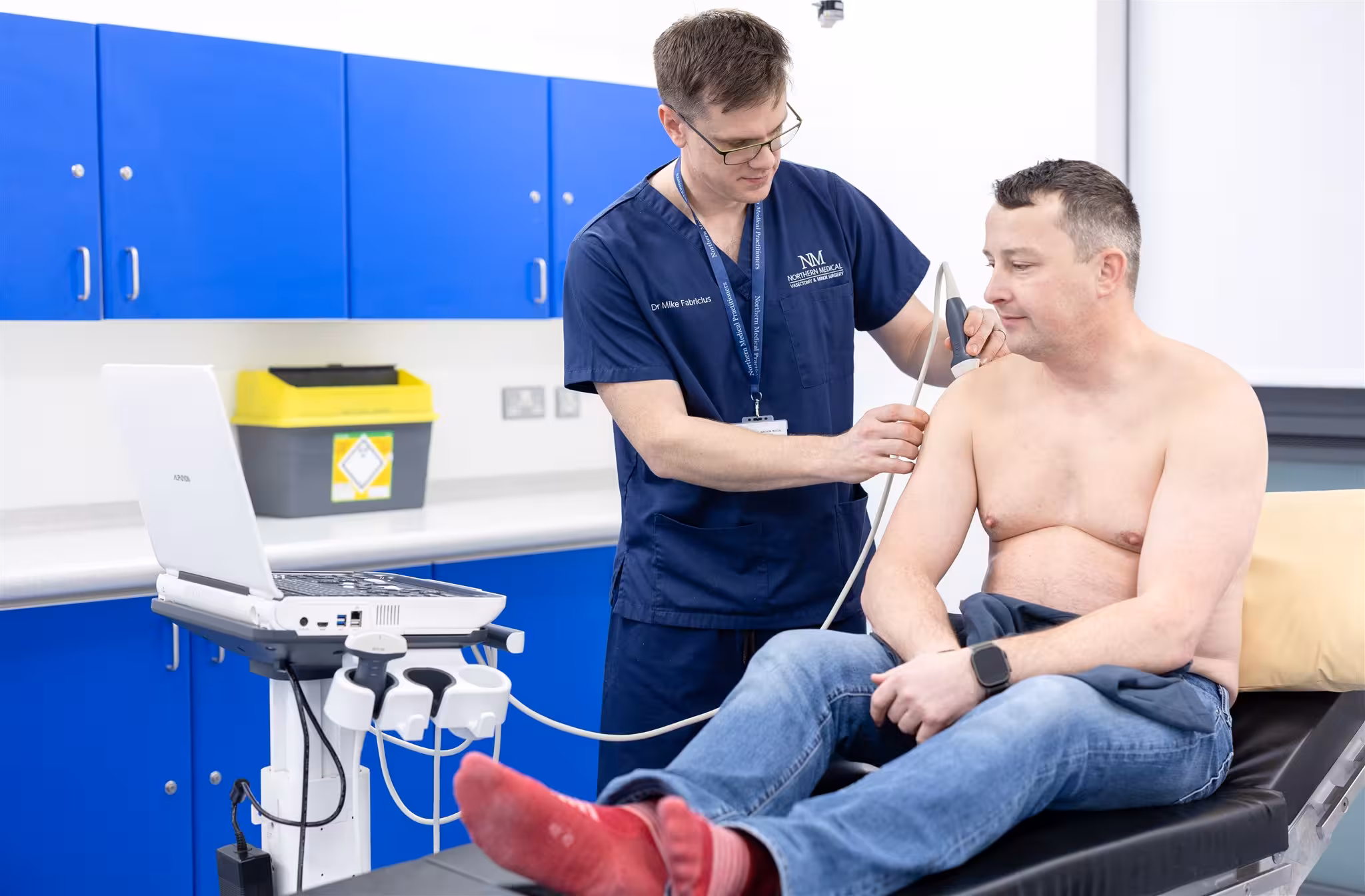 Dr Mike Fabricius of Northern Medical Practitioners performing a diagnostic ultrasound scan on a male patient's bare shoulder in a clinic room.