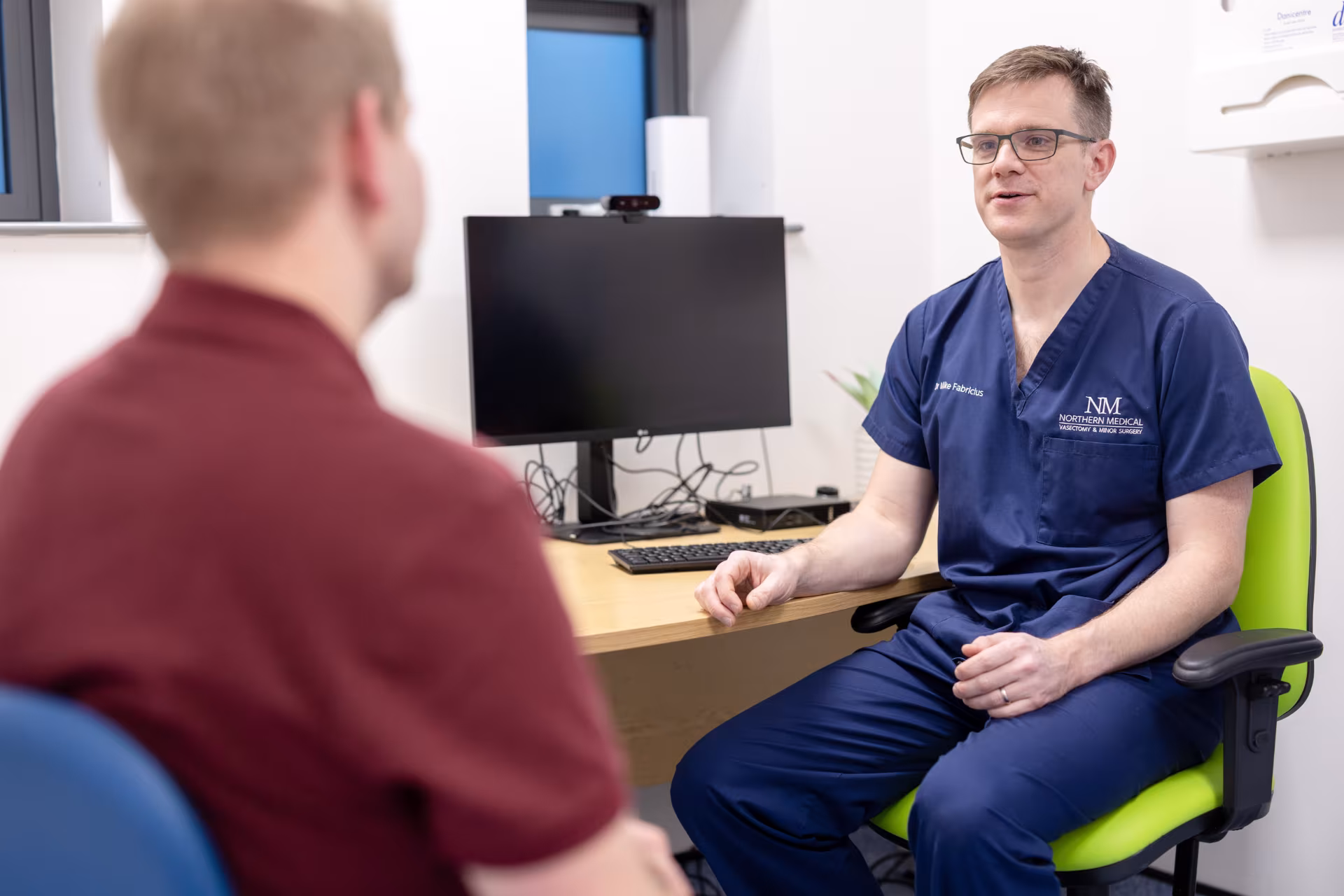 Dr Mike Fabricius of Northern Medical Practitioners, wearing blue scrubs and glasses, speaking with a patient during a consultation at his clinic desk.
